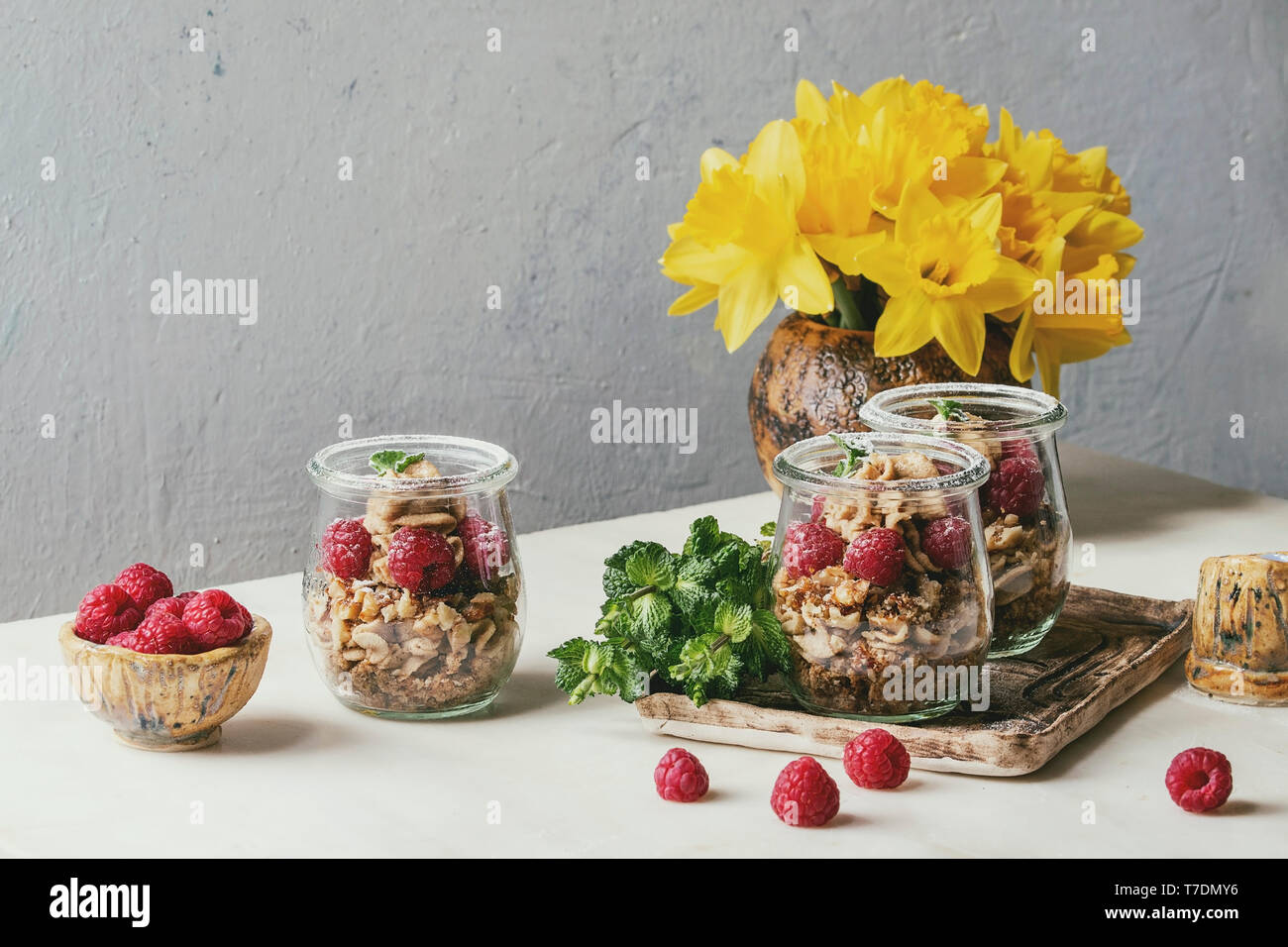 Layered dessert in jars. Biscuit, coffee cream, nuts, raspberries, fresh mint. Served on ceramic tray with daffodils flowers, berries and greens over Stock Photo