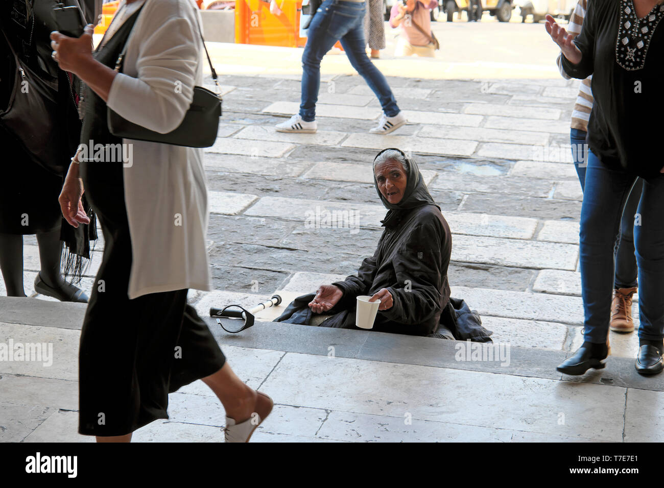 Poor old woman sitting on steps of a church holding out a cup and begging for money while people walk past in Lisbon Portugal Europe EU  KATHY DEWITT Stock Photo