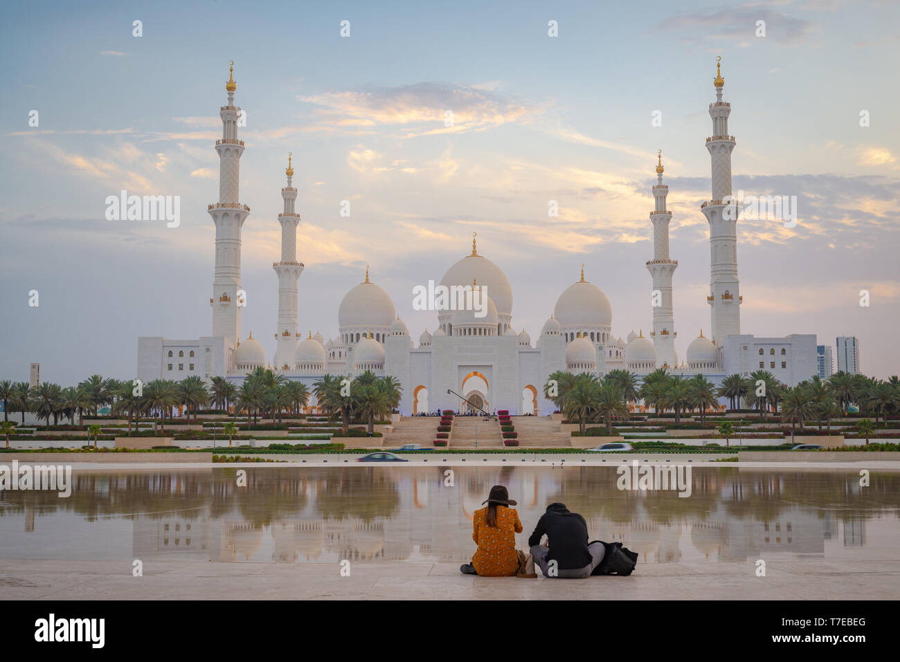 Axial view of the Great Mosque of Abu Dhabi at sunset with reflection over a water mirror with a couple seen from behind, United Arab Emirate Stock Photo