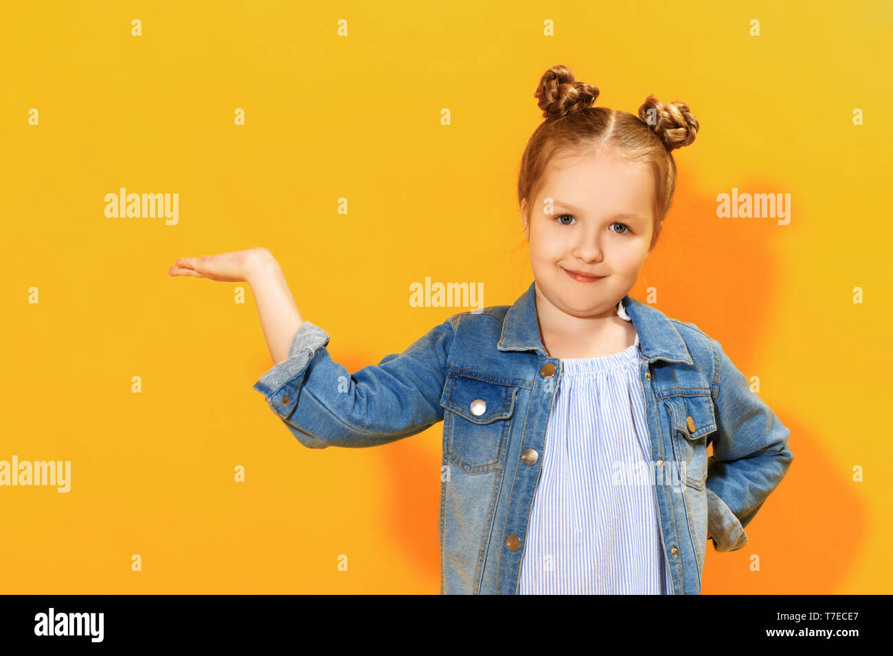 Closeup portrait of a cute attractive little girl on yellow background. The child holds in the palm a copy of the empty blank space and looks into the Stock Photo