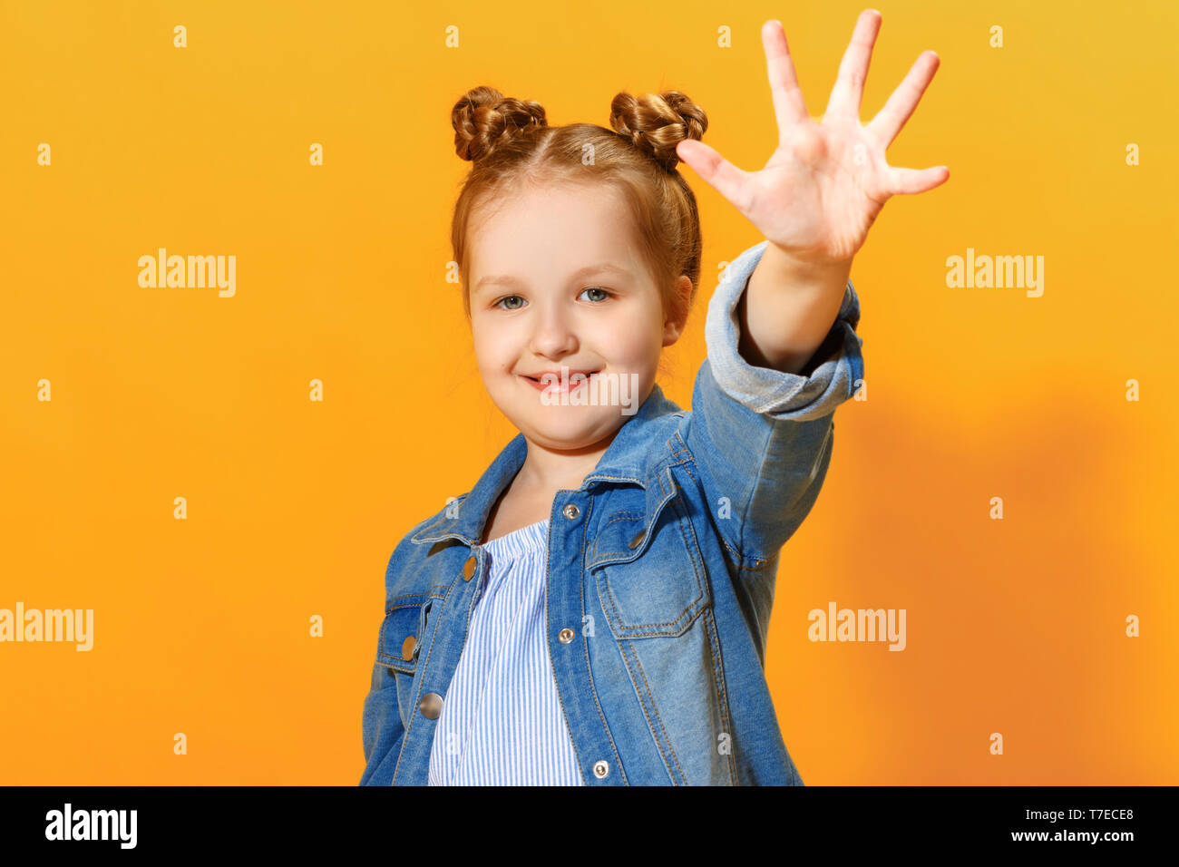 Portrait of a funny little girl on a yellow background. Preschool child shows the palm, give five. Stock Photo