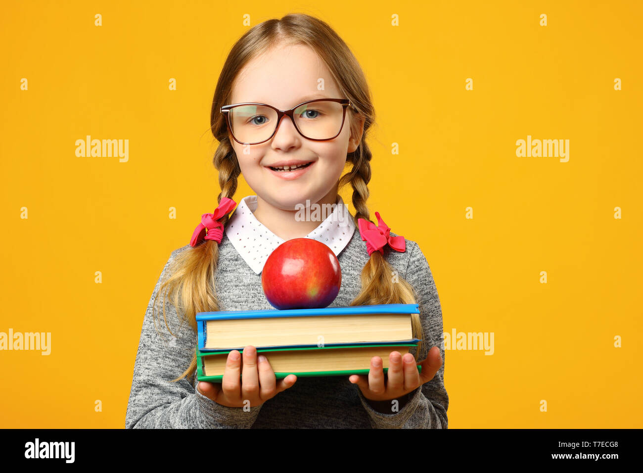 Portrait of a cheerful little child girl in glasses on yellow background. Schoolgirl is holding a stack of books and an apple. The concept of educatio Stock Photo