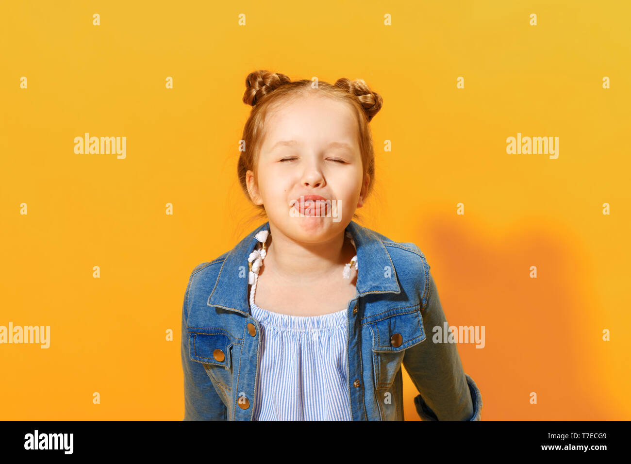 Cheerful little child girl closed her eyes and shows tongue. Yellow background, studio. Stock Photo