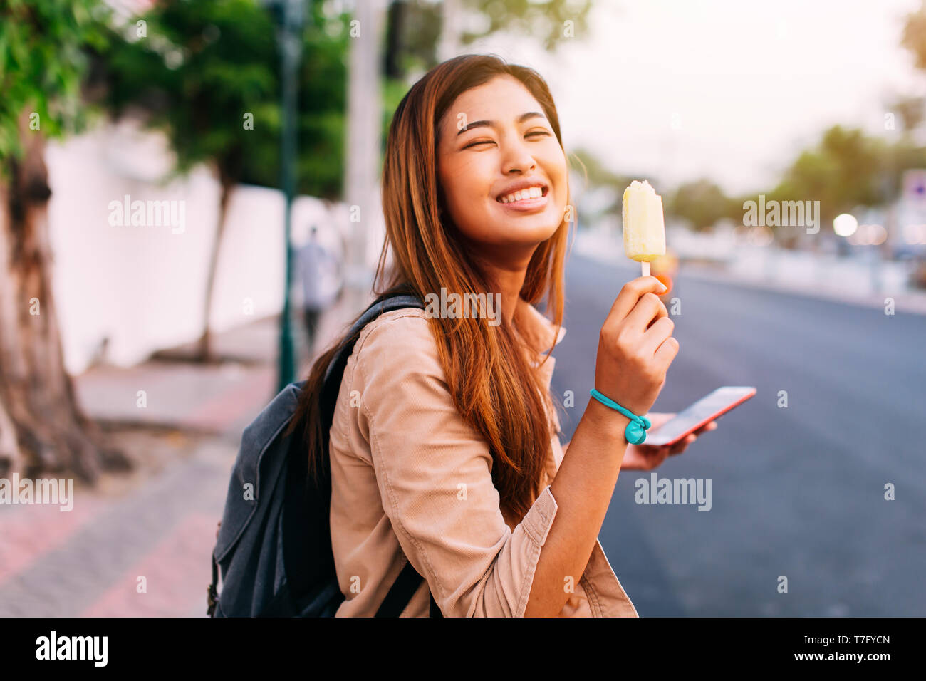 Side view of charming Asian woman holding smartphone and eating ice-cream while smiling at camera on sunny street Stock Photo