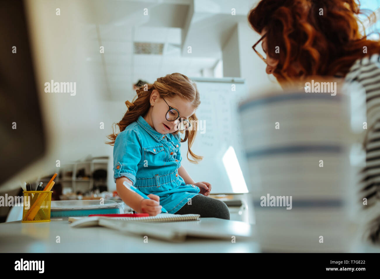 Girl wearing denim dress painting on working table of mother Stock Photo