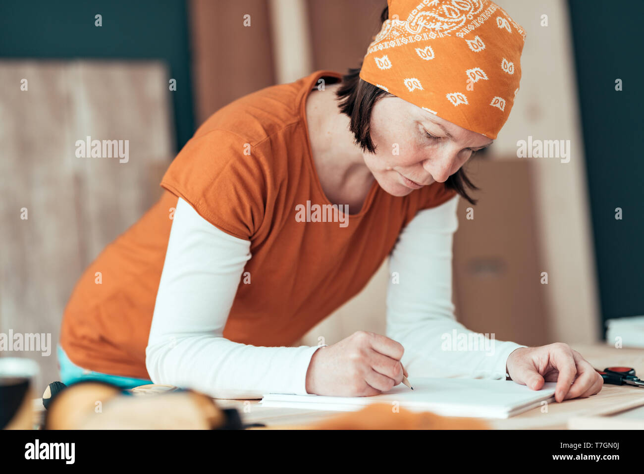 Female carpenter wearing bandanna is sketching project draft on clipboard note paper in her small business woodwork workshop Stock Photo