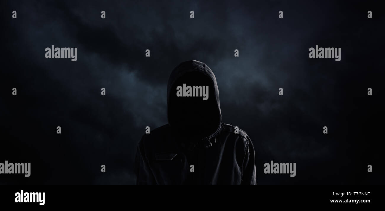 Spooky hooded person with obscured face against the dark dramatic sky Stock Photo