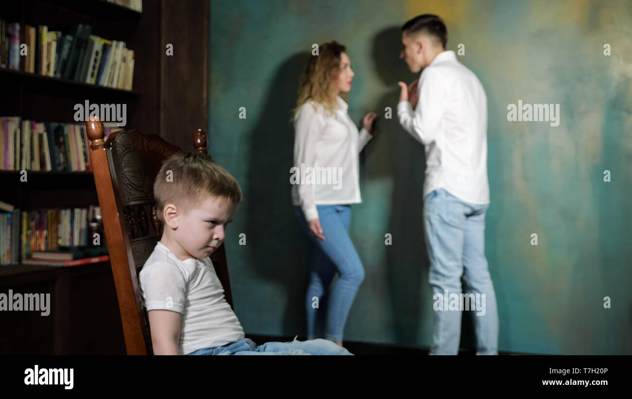 Couple of parents arguing in front of their kid. Sad little boy is listening his parent's shouting on each other, sitting on the chair, side view. Stock Photo