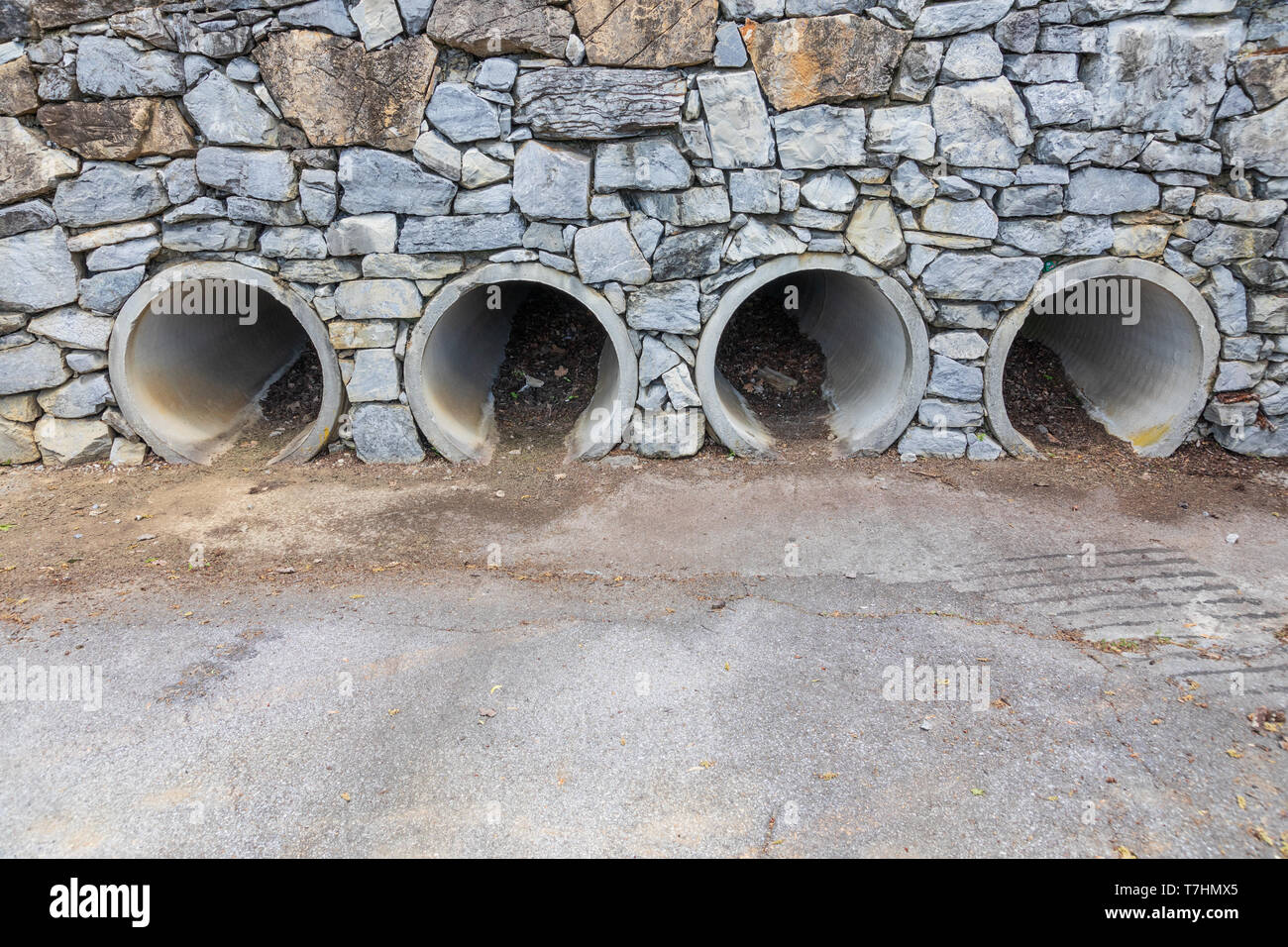 Four large pipes functioning as culverts through a rock wall, opening onto concrete parking lot. Stock Photo