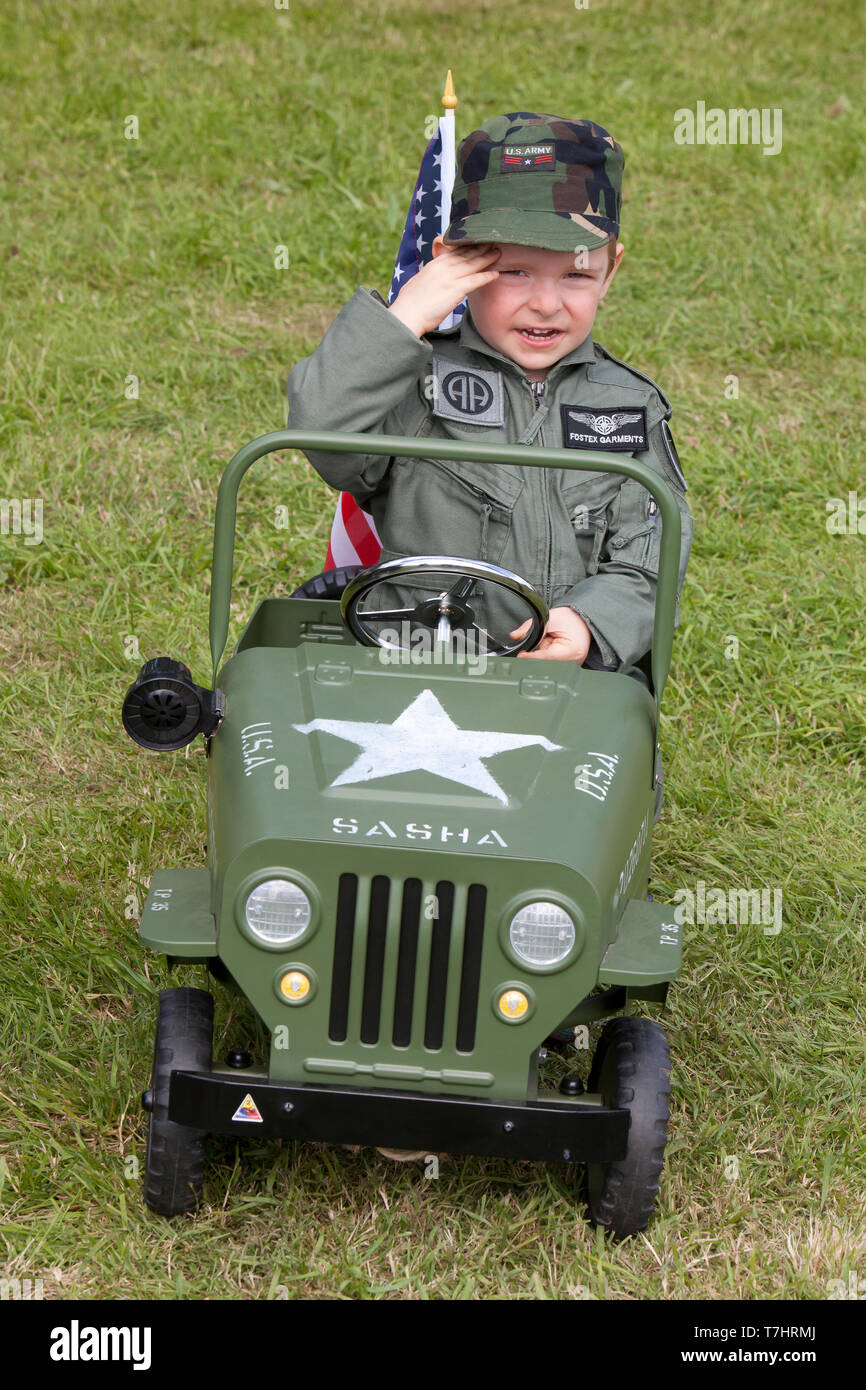 A young boy dressed in an overall of the 82nd Airborne Division saluting whilst driving a Willys jeep at the D-Day celebrations in Normandy, France Stock Photo