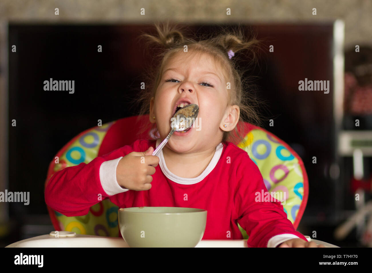 Portrait of little girl licking spoon eating porridge sitting in a feeding chair Stock Photo
