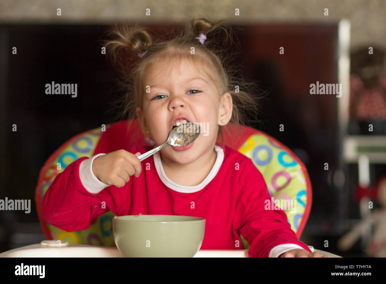 Portrait of little girl licking spoon eating porridge sitting in a feeding chair Stock Photo