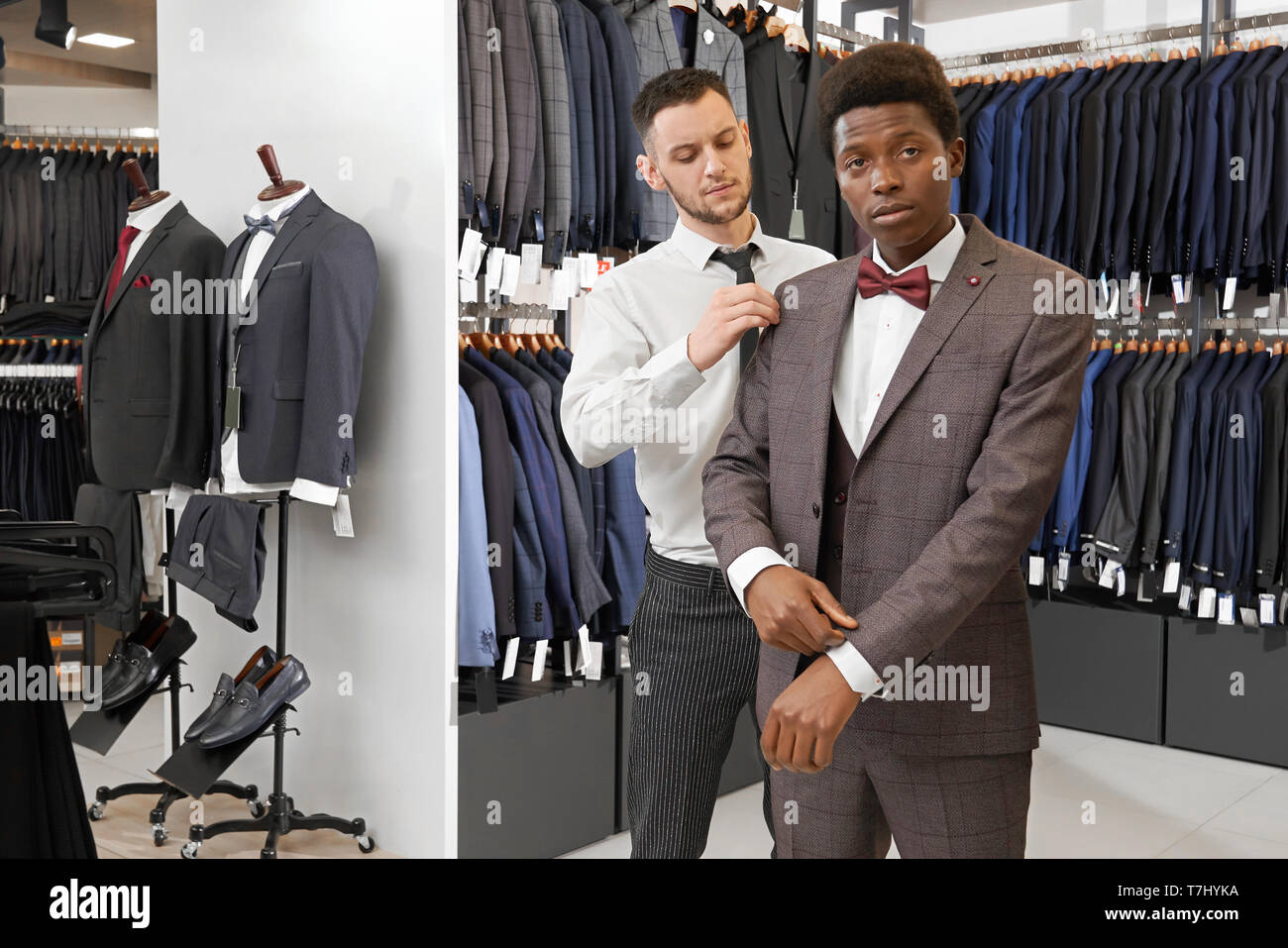 Assistant of fashionable boutique trying on and choosing suit for young client. Man in white shirt and suit looking at camera. Stylish showroom with clothing for successful men. Stock Photo