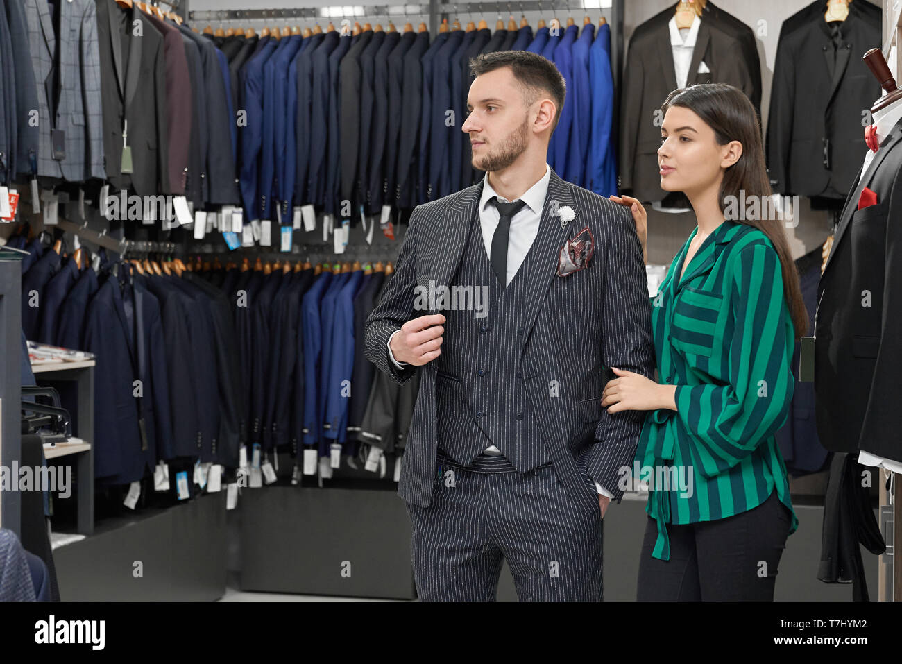 Handsome gentleman in fashionable grey striped suit and black necktie, white shirt posing in store, looking away. Pretty girl shop assistant helping client with choosing clothing. Stock Photo