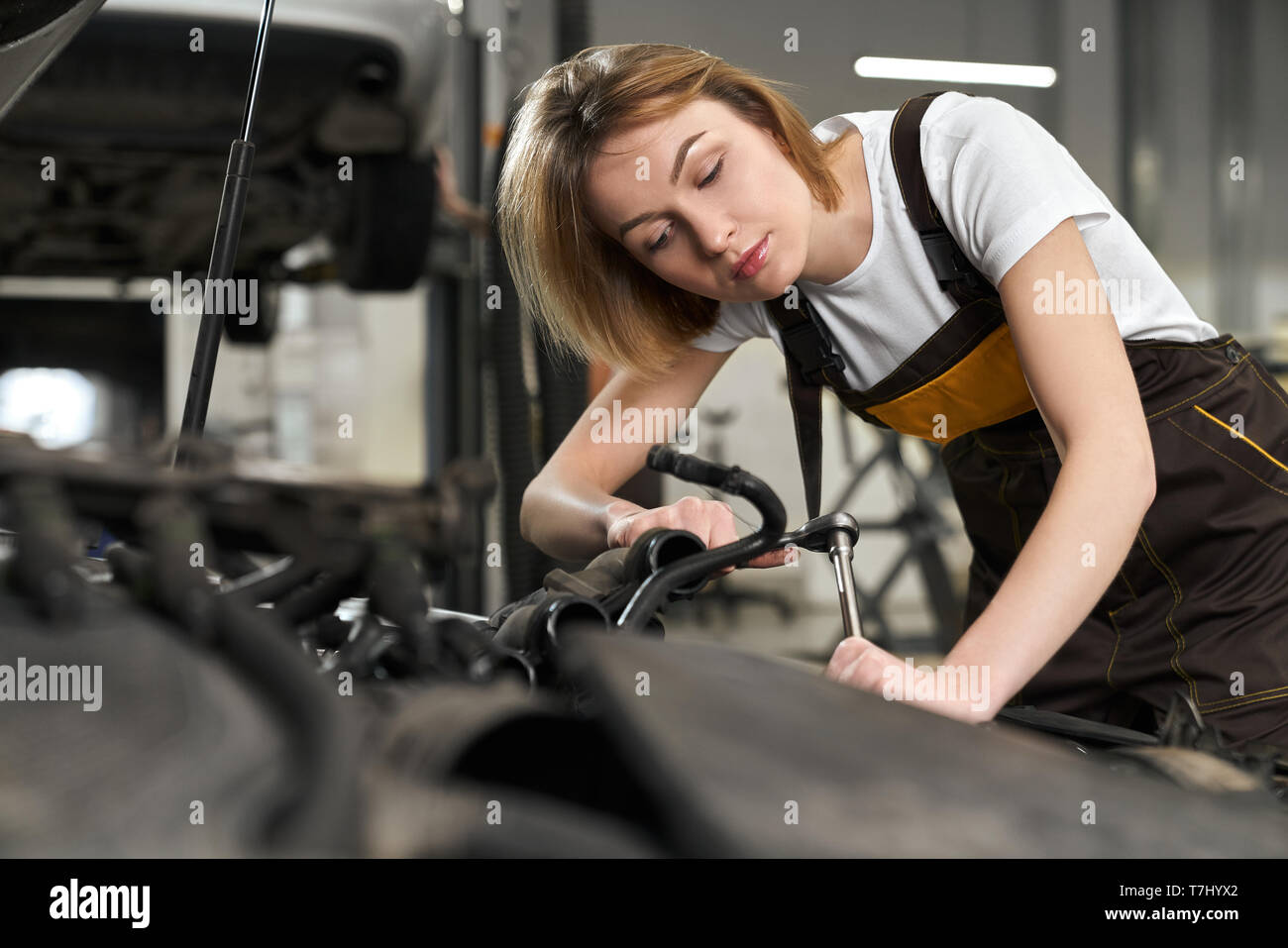 Pretty young girl in uniform working in autoservice station fixing and repairing car. Beautiful female mechanic with blonde short hair holding tool, wench and looking at car motor. Stock Photo