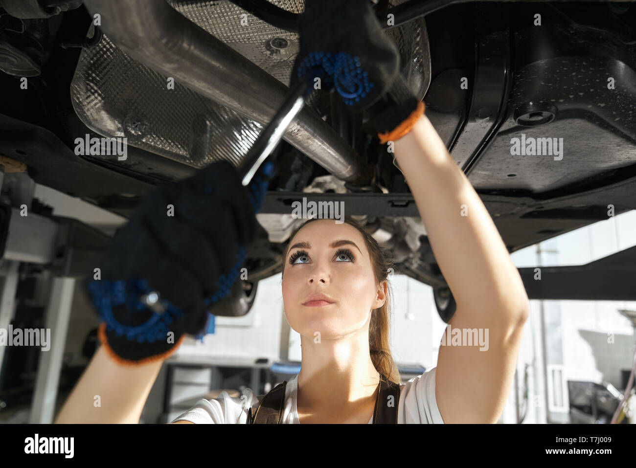 Close up of beautiful young woman fixing undercarriage of automobile. Concentrated girl in black gloves holding wrench, looking up on vehicle lifted on bridge. Autoservice station. Stock Photo