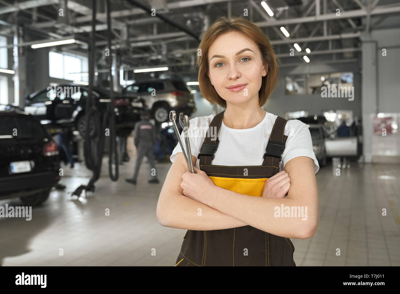 Beautiful, handsome female mechanic looking at camera, posing, standing in autoservice. Pretty young woman wearing in white t shirt and coveralls, holding wrenches. Stock Photo