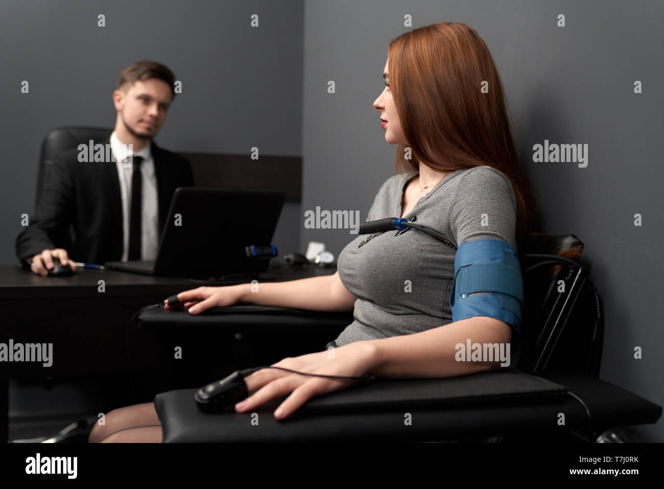 Woman sitting in chair with attached finger sensors and indicators, looking at lie detector. Young man at black table, testing woman with computer polygraph. Stock Photo