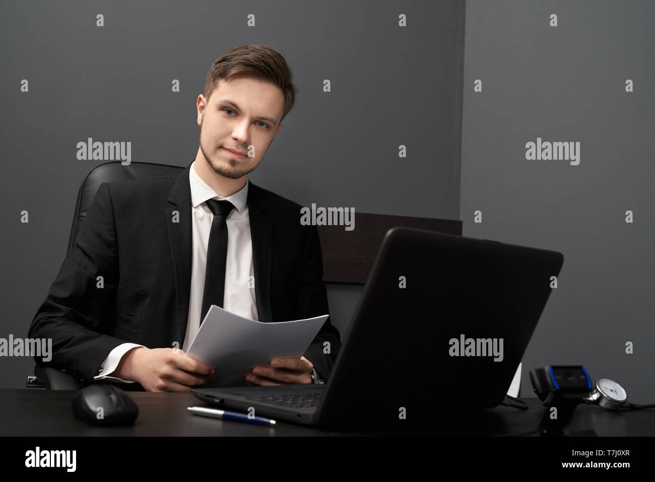Young man sitting at black table with computer polygraph in grey room. Handsome man with beard in black suit holding paper, looking at camera. Concept of lie detector. Stock Photo