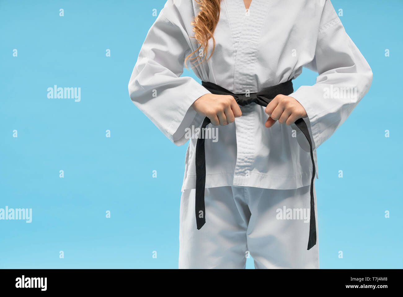 Front view of white kimono on little fighter keeping black belt on blue isolated background. Sporty child standing and posing in studio. Strong girl doing karate and judo. Concept of sport. Stock Photo