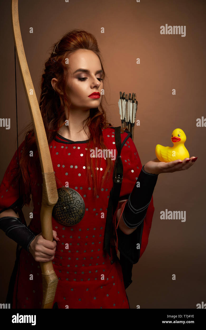 Beautiful woman warrior wearing in medieval red tunic holding small, yellow rubber duck on hand, looking at it. Serious model with red lips and ginger hair posing, holding bow. Stock Photo