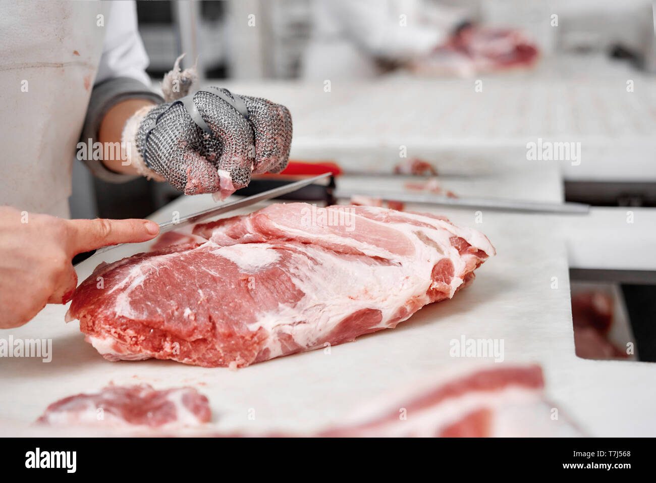 Close up of big fresh chopped pieces of meat and male hands of worker in special gloves. Butcher holding knife and cutting meat pork or beef on table. Manufacturing of raw meat. Stock Photo