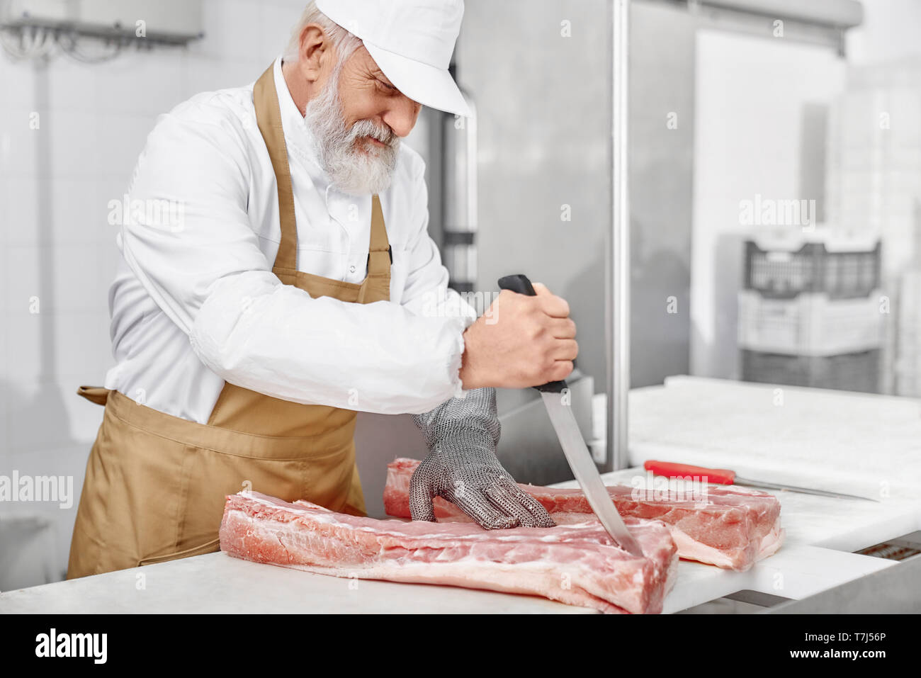 https://c8.alamy.com/comp/T7J56P/elderly-man-working-on-meat-production-professional-butcher-in-white-uniform-cap-brown-apron-and-special-gloves-standing-near-table-holding-knife-and-cutting-meat-food-industry-T7J56P.jpg