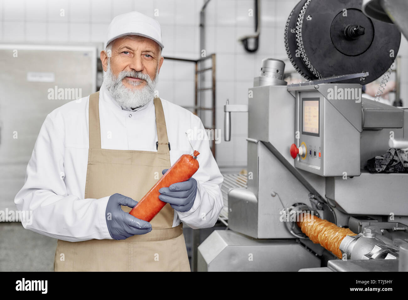 Handsome worker standing near modern new equipment for sausage production, holding sausage, looking at camera, posing. Food industry. Elderly man wearing in white uniform and apron. Stock Photo