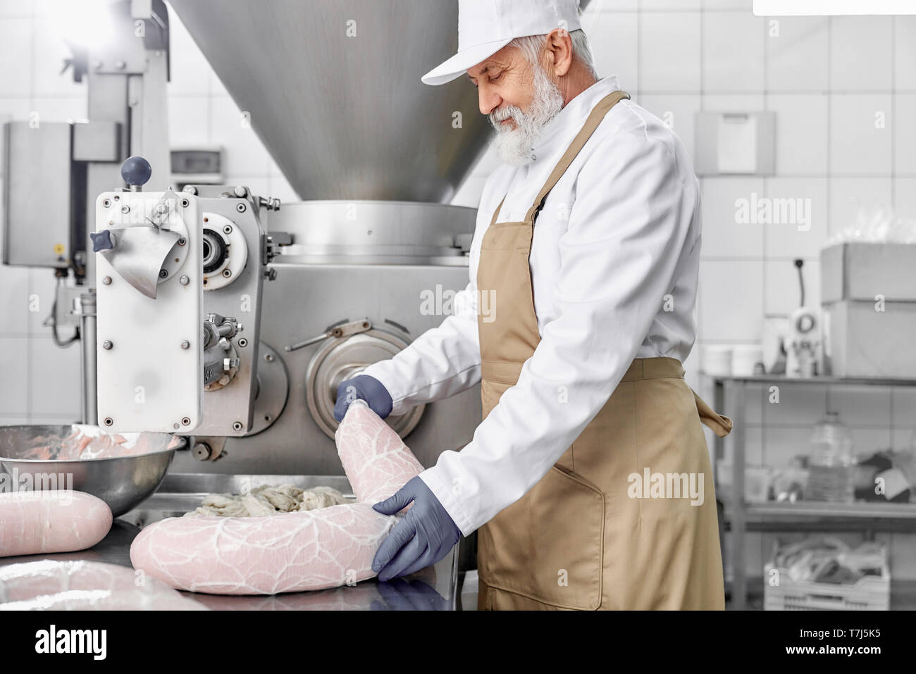 Butcher man standing and working on equipment for production of sassages.  Elderly, handsome man in white uniform and brown apron, rubber gloves  checking and observing Stock Photo - Alamy