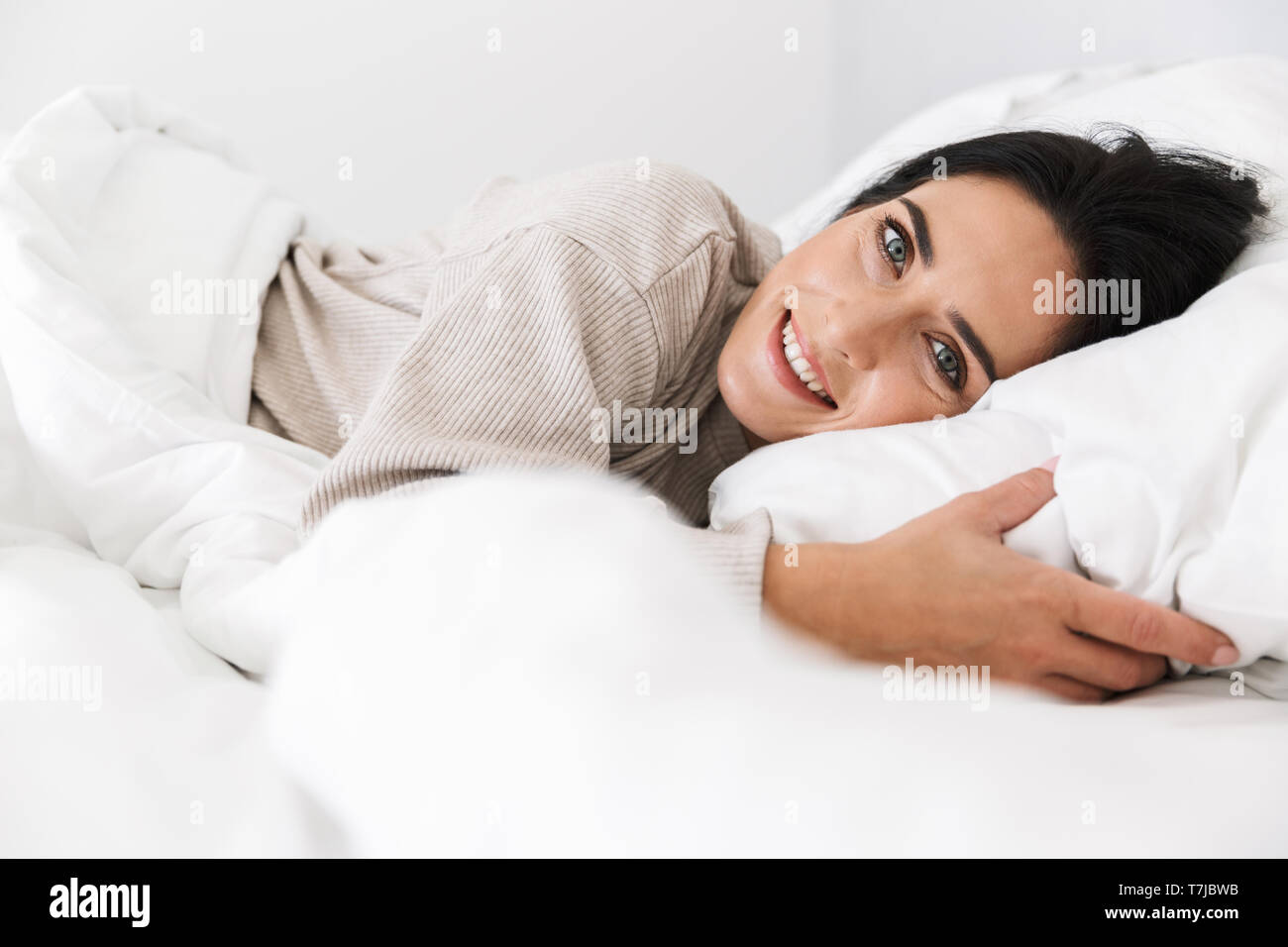 Photo of joyful woman 30s smiling while lying in bed with white linen at home Stock Photo