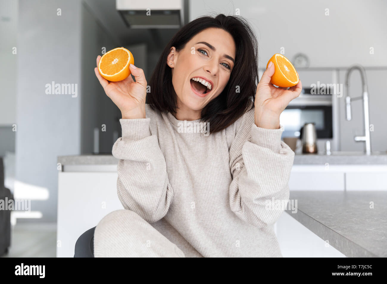 Image of joyful woman 30s holding two pieces of orange while sitting in living room Stock Photo