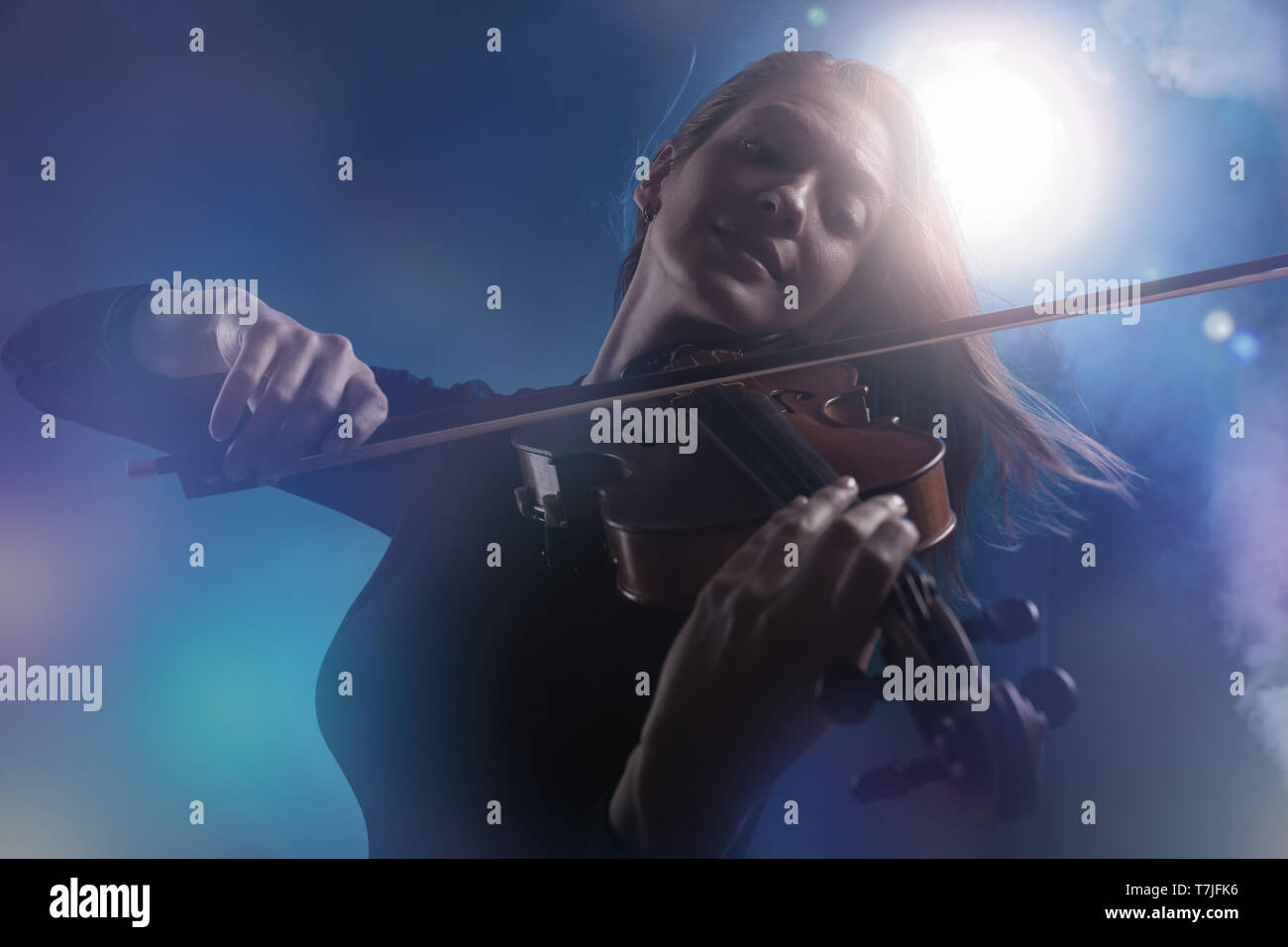Beautiful young woman playing the violin on dark blue background. Fog in the background. Studio shot Stock Photo