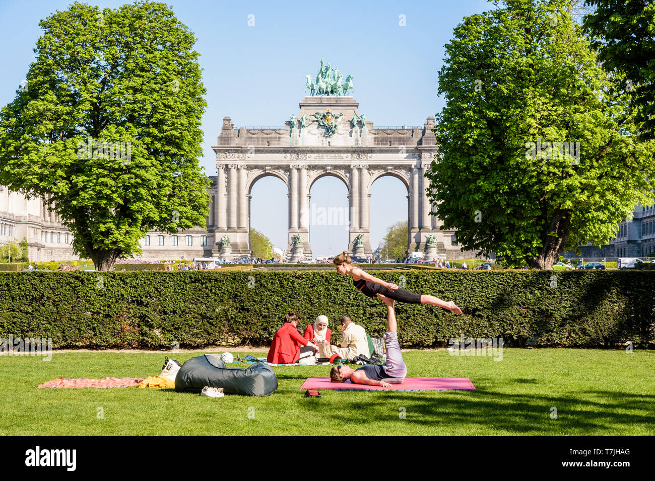 A young couple is practicing acroyoga on the grass in the Cinquantenaire park in Brussels, Belgium, with the arcade du Cinquantenaire in the distance. Stock Photo