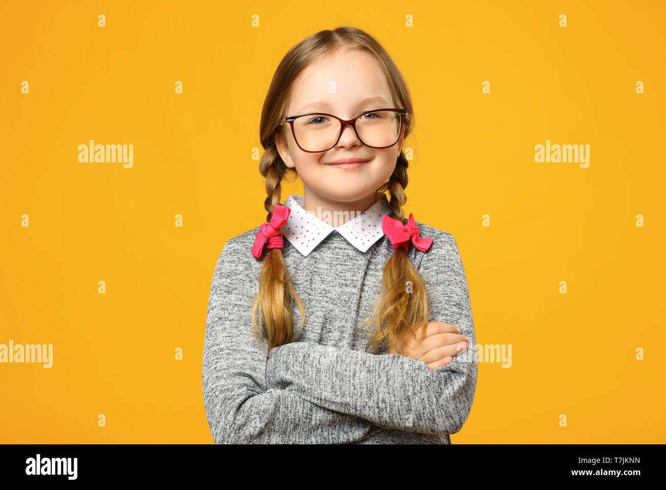 Closeup portrait of a funny little girl in glasses on a yellow background. Child schoolgirl folded her arms and looks into the camera. Stock Photo