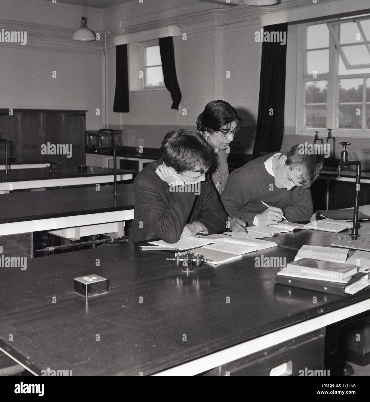 1960s, historical, in an empty secondary school classroom, a female mathematics teacher overseeing the study or book work of two male pupils, who are doing extra study or revision after class, South London, England, UK. Stock Photo