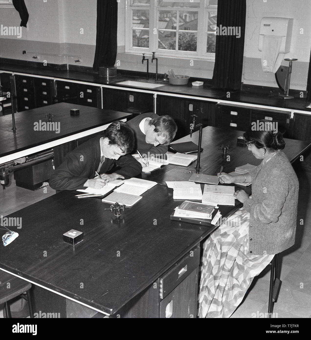 1960s, historical, in an empty secondary school classroom, a female mathematics teacher sitting at a desk overseeing two male pupils at the same desk doing additional study work after class, South London, England, UK. Stock Photo