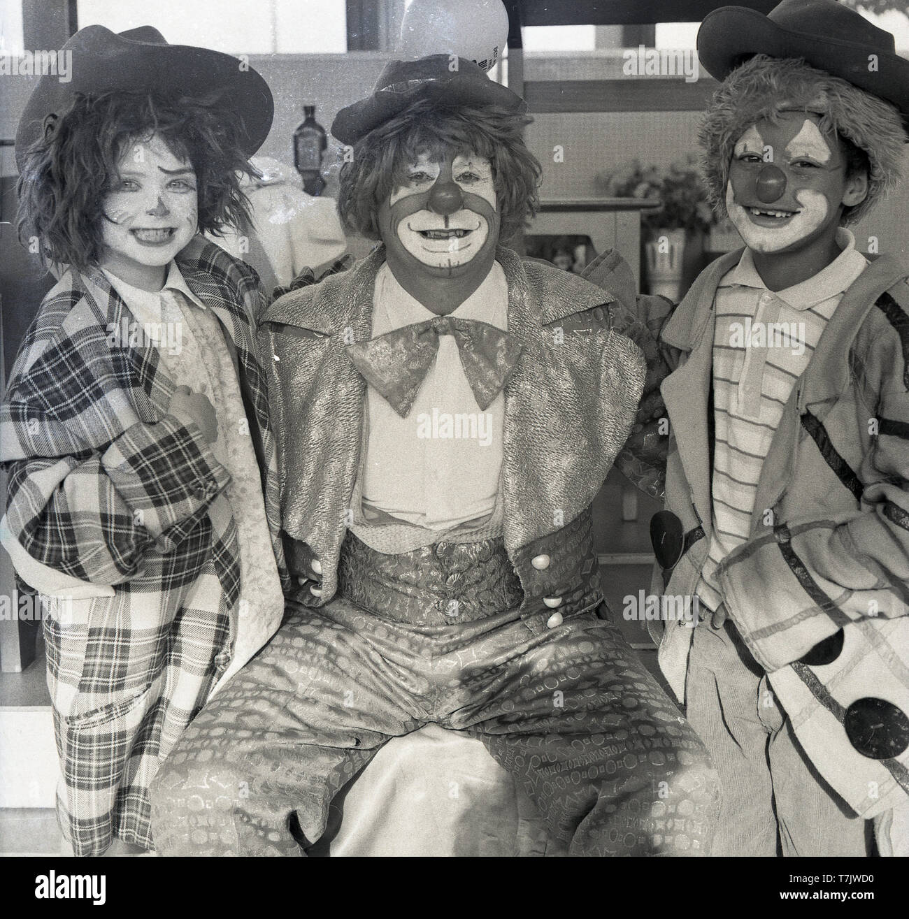 1960s, historical, a circus clown visiting a primary school sitting beside with young school children both wearing fancy dress or funny outfits and with their faces painted as a clown, England, UK. Stock Photo