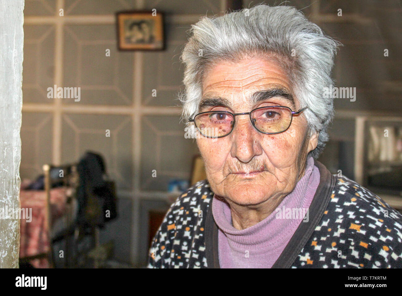 An elderly woman with glasses looks into the lens of the photographer behind her photo on the wall Stock Photo