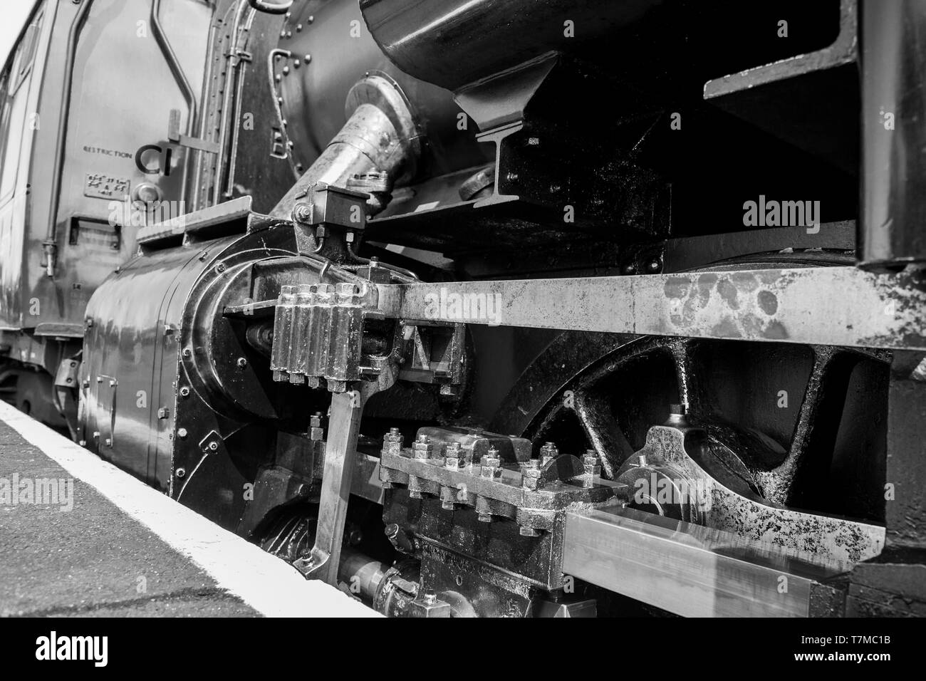 Low angle, black and white close up of vintage UK steam locomotive driving mechanism, as steam train is stationary alongside station platform. Stock Photo
