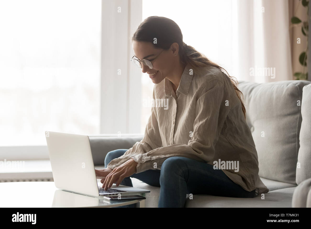 Woman in glasses sitting on couch smiling typing on computer Stock Photo