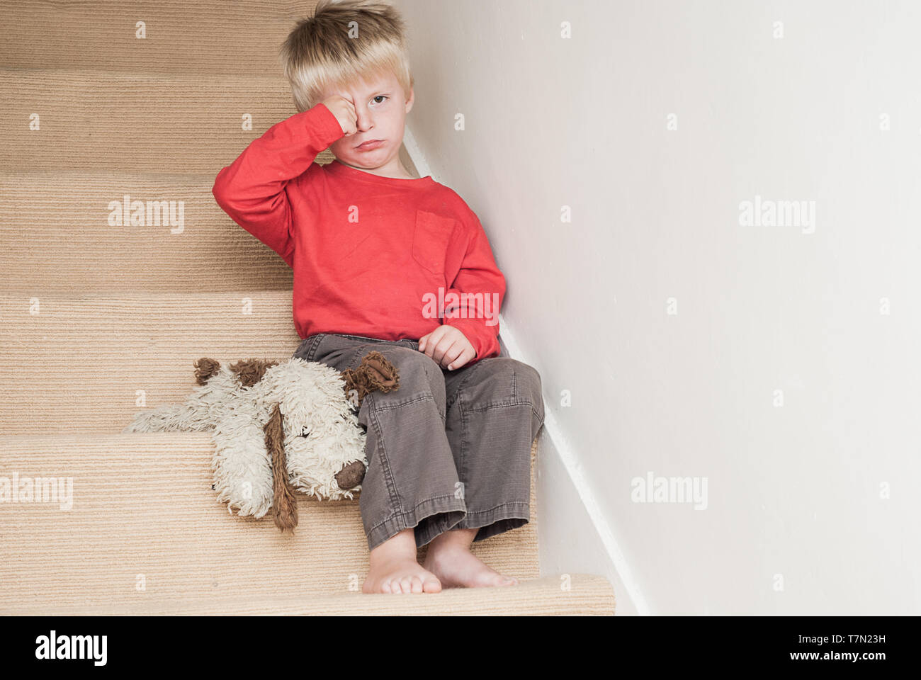 Lonely upset child sitting on the stairs in the home with his teddy next to him. Stock Photo