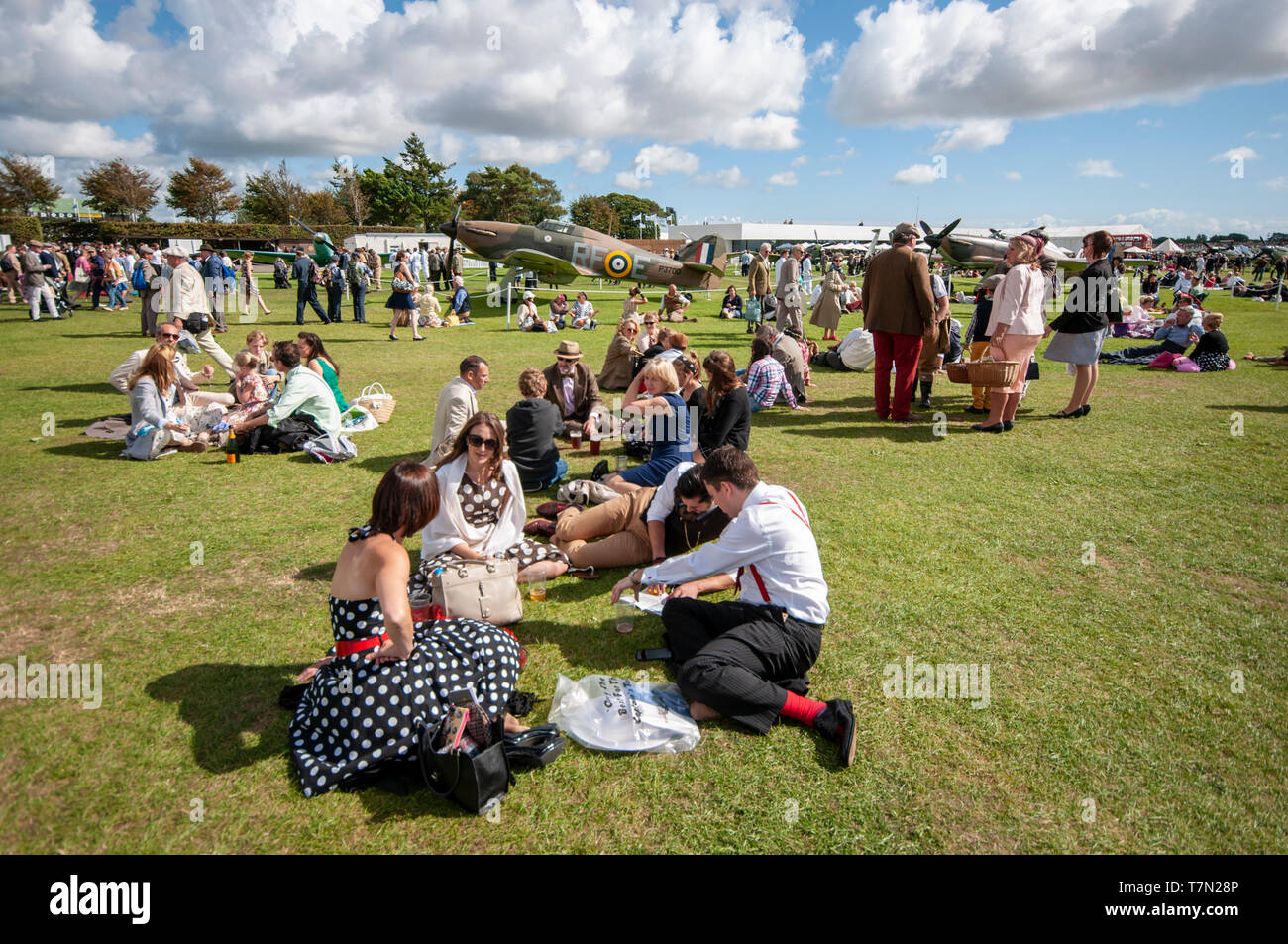 People in period attire at the Goodwood Revival, UK. Picnics on the lawn with Second World War planes. Period dress, costumes. British Stock Photo