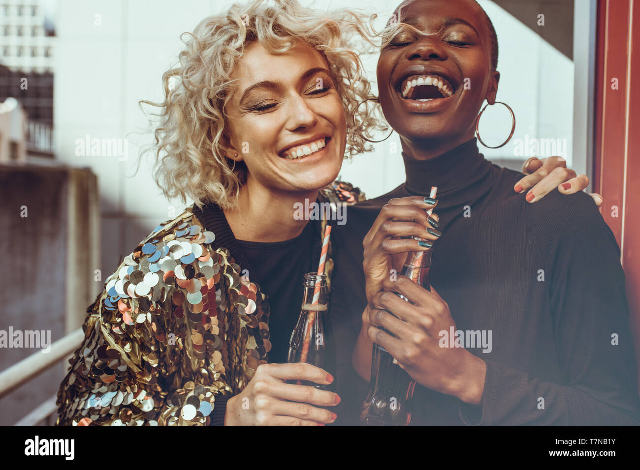 Multi-ethnic women friends standing together with cola and laughing. Two young female friends hanging out on their weekend. Stock Photo