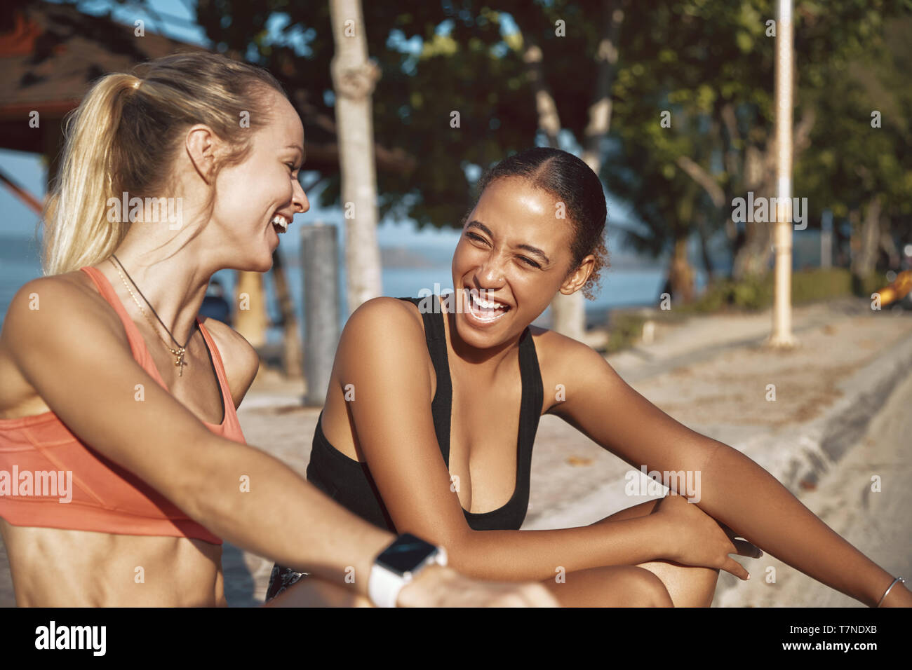 Two fit young women in sportswear stting on a curb talking and laughing while taking a break from their run Stock Photo