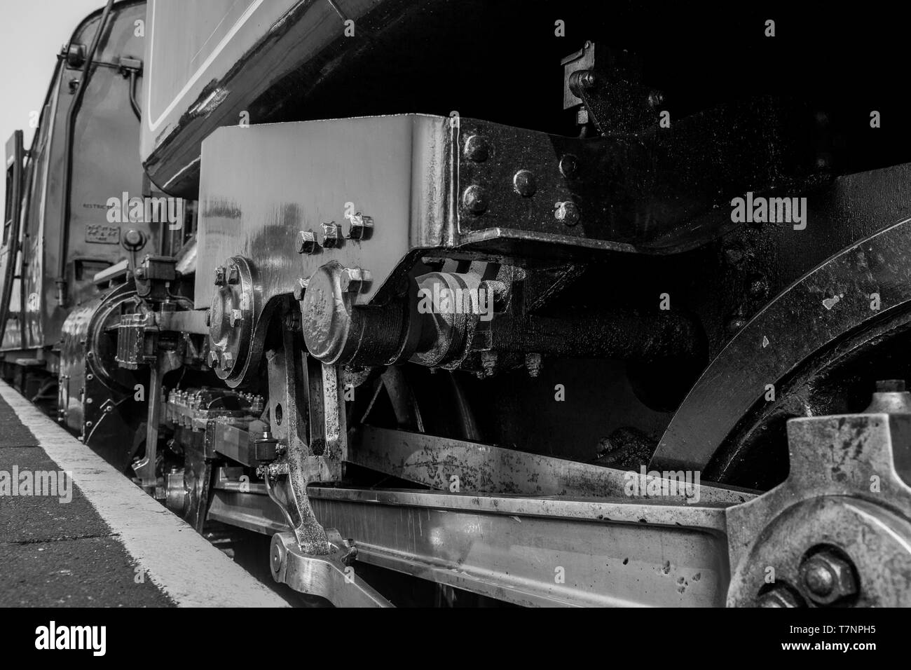Low angle, black and white close up of vintage UK steam locomotive driving mechanism, as steam train is stationary alongside station platform. Stock Photo