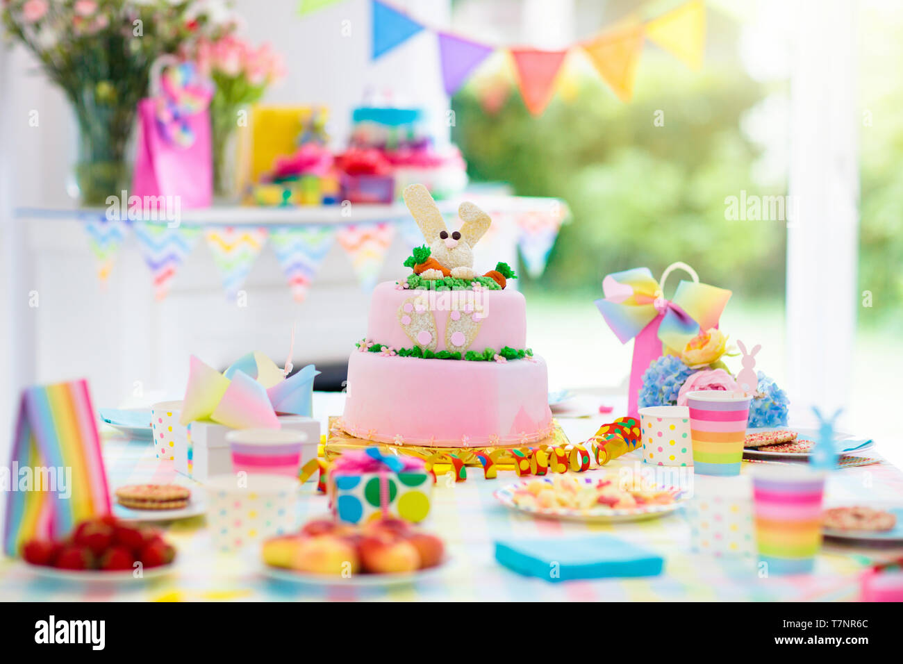 Kids birthday party decoration and cake. Decorated table for child birthday celebration. Rainbow bunny cake for little girl. Room with festive balloon Stock Photo