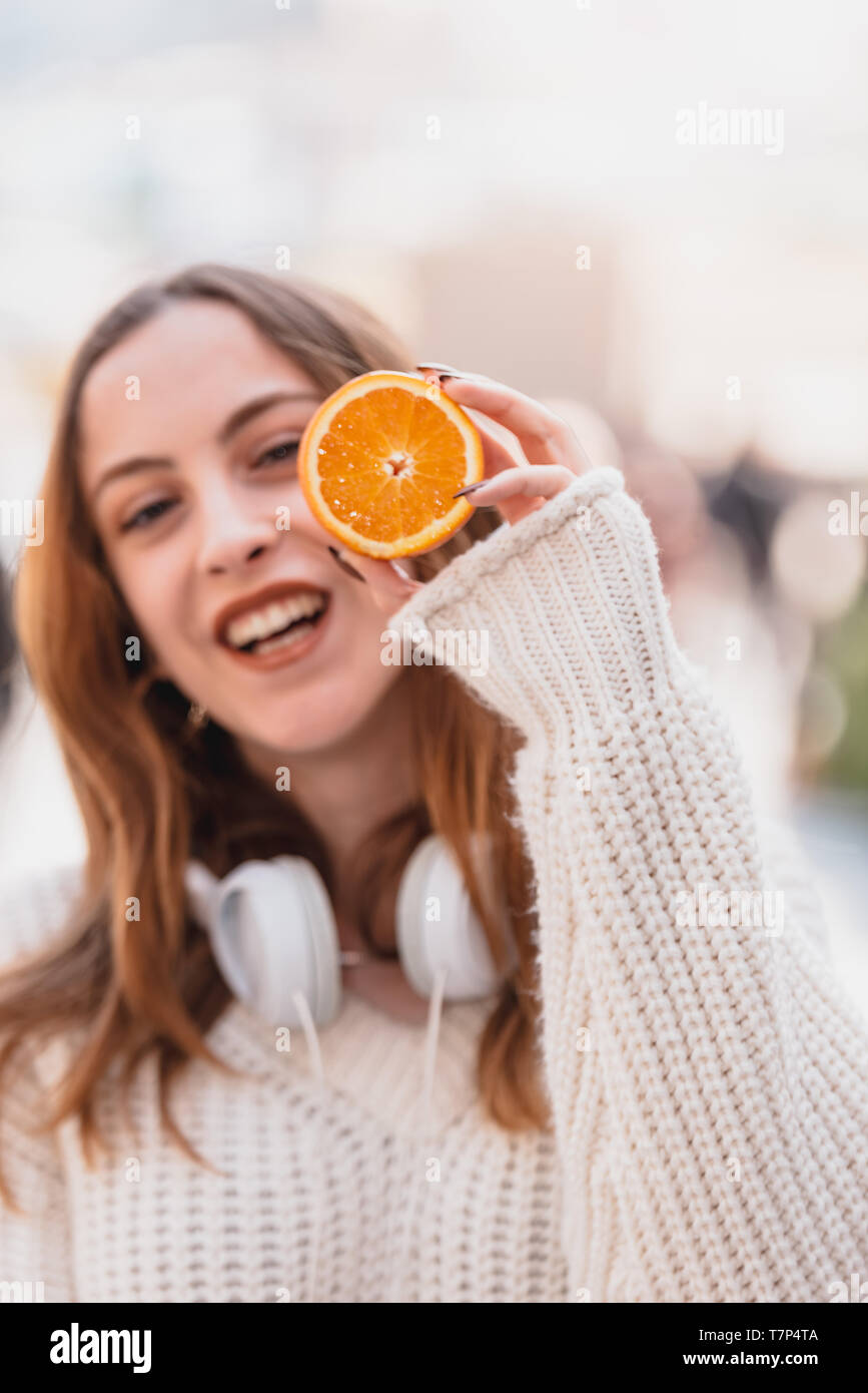 Portrait of beautiful attractive young trendy girl with headphones holds half of citrus fruit in hand, covering her eye Stock Photo