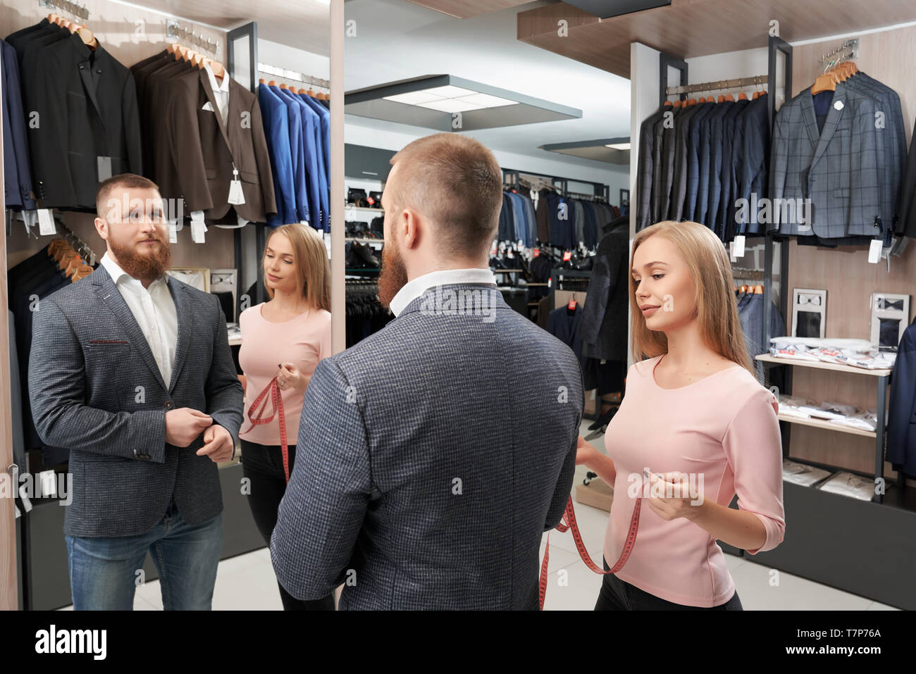 View from side of bearded man trying new white shirt and grey casual jacket in shop. Smiling man standing in front of mirror while female tailor measuring length of sleeve. Concept of fitting. Stock Photo