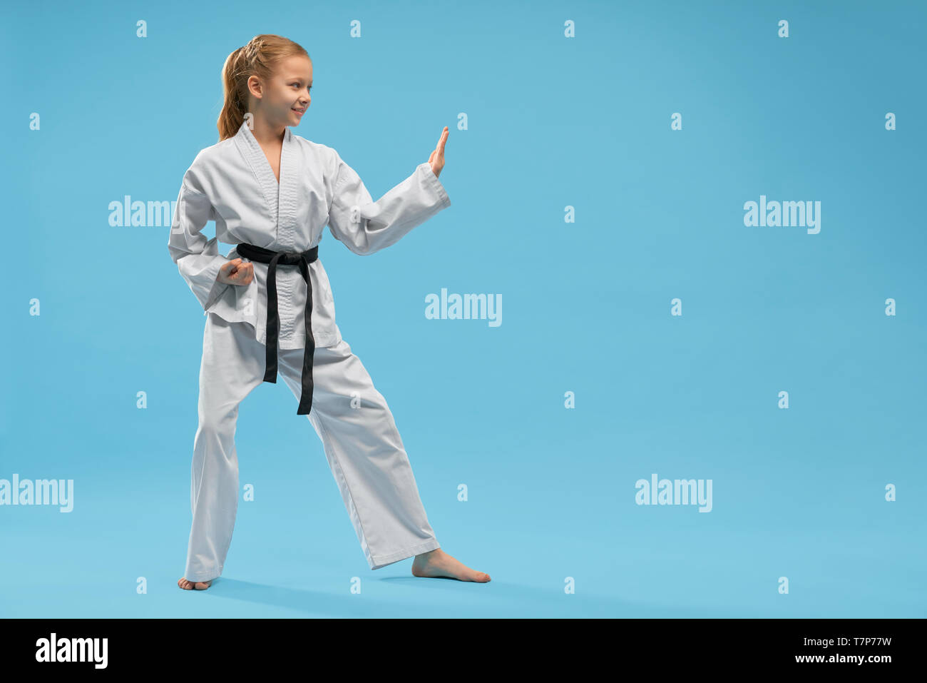 Side view of smiling girl wearing white kimono looking forward and training karate on blue isolated background. Active child doing sport and practising taekwondo. Concept of martial arts and combat. Stock Photo