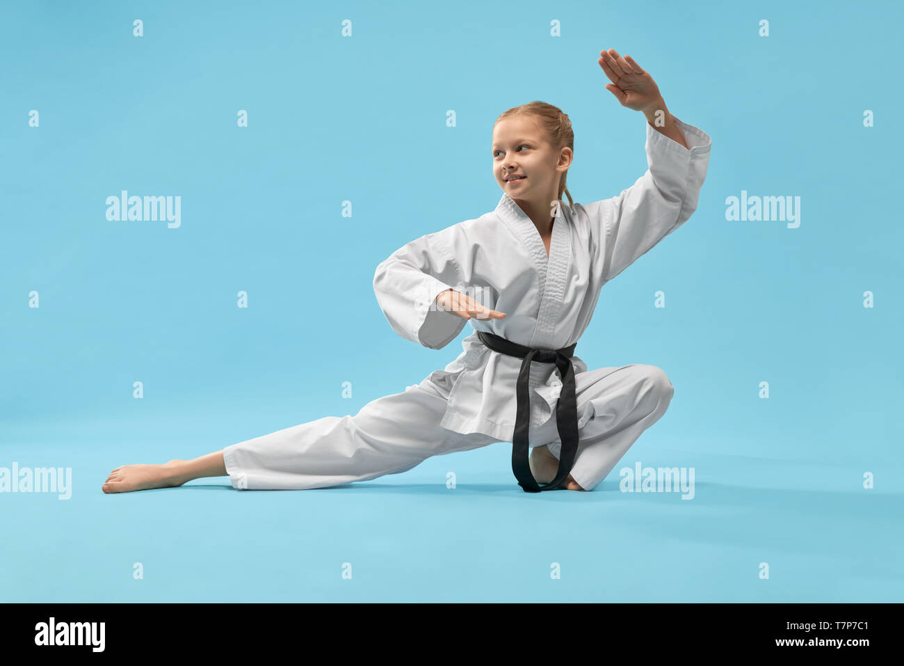 Cheerful little sports woman sitting on twine and practising karate on blue isolated background. Active girl wearing white kimono and black belt doing sport in studio. Concept of karate and jujitsu. Stock Photo